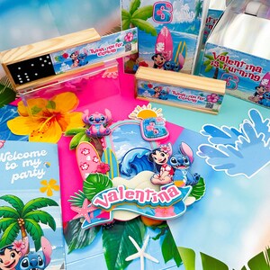 May include: Party decorations with Lilo and Stitch theme. Includes a cake topper, invitation cards, gift boxes, and wooden boxes with dominoes. The theme is a beach party with surfboards and tropical flowers. The text "Valentina is turning 6" is visible.