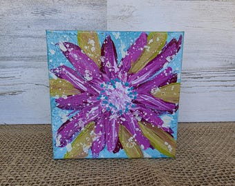 以下が含まれることがあります： 鮮やかな抽象的な花の絵が描かれた正方形のキャンバスアート。このアートワークは、明るい青色の背景に、黄緑色の花びらを持つ大きな紫色の花を特徴としています。白いペイントのスプラッシュがテクスチャを追加します。