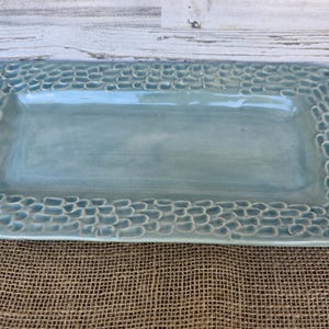 May include: A rectangular, light blue ceramic serving platter. The edges feature a textured, pebble-like design. The platter is displayed on a burlap surface, with a white wooden background.