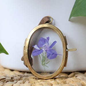 May include: An oval-shaped brass frame with a hinged side closure, containing a pressed purple flower behind clear glass. A brown suede cord is attached to the top loop of the metal frame, which features a metallic gold-toned finish.