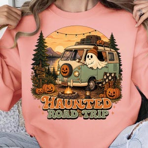May include: A peach-coloured sweatshirt featuring a graphic of a light green vintage van with a ghost in a beanie, a campfire, pine trees, and pumpkins. The text reads HAUNTED ROAD TRIP in orange and green letters against a sunset background.