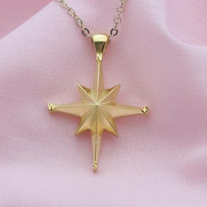 Könnte beinhalten: Eine goldfarbene Stern-Anhänger-Halskette. Der Anhänger hat acht Spitzen und hängt an einer zarten Kette. Die Halskette wird auf einem weichen, rosa Stoffhintergrund präsentiert.