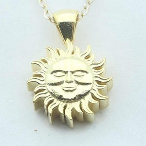 May include: A gold-colored pendant necklace featuring a sun design. The sun pendant has a smiling face and radiating rays. The necklace chain is also gold-colored and hangs against a white background.