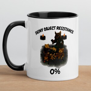 Puede incluir: Taza de cerámica blanca con interior y asa negros. La taza presenta el texto "SHINY OBJECT RESISTANCE" y una ilustración de dibujos animados de una figura oscura y pequeñas criaturas. El número "0%" está debajo de la ilustración.