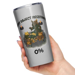 Puede incluir: Un vaso plateado con tapa negra. Presenta una ilustración de dibujos animados de una figura encapuchada, velas y pequeñas criaturas. También se ve el texto "ANY OBJECT RESISTANCE" y "0%".