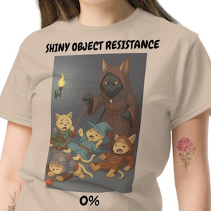Puede incluir: Camiseta beige con el texto "SHINY OBJECT RESISTANCE" sobre una ilustración de dibujos animados de personajes felinos en una mazmorra. La ilustración incluye una figura encapuchada y el número "0%" en la parte inferior.