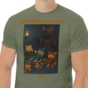 Puede incluir: Camiseta verde oliva con una ilustración de dibujos animados. La imagen muestra personajes con forma de gato en una mazmorra, con el texto "SHINY OBJECT RESISTANCE" y "0%". Una figura controla a otras con un láser rojo.