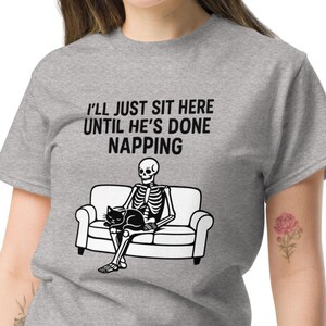 May include: A gray t-shirt with the text "I'LL JUST SIT HERE UNTIL HE'S DONE NAPPING." The shirt features a black and white graphic of a skeleton sitting on a couch with a black cat.