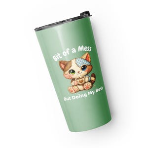 May include: A light green tumbler with a black lid featuring a cartoon cat illustration. The cat is holding a cup and the text reads "Bit of a Mess But Doing My Best". The tumbler is designed for beverages.
