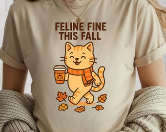 Könnte beinhalten: Beiges T-Shirt mit dem Text "Feline Fine This Fall" und einer Cartoon-Katze. Die Katze, mit Schal, hält einen Pumpkin Spice Latte und ist von Herbstblättern umgeben.