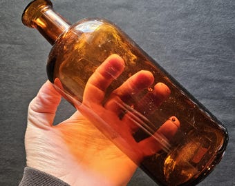 May include: A brown glass bottle with a long neck and rounded body. The bottle is held in a hand, and the light shines through the glass, highlighting its amber color. The bottle appears to be vintage or antique.