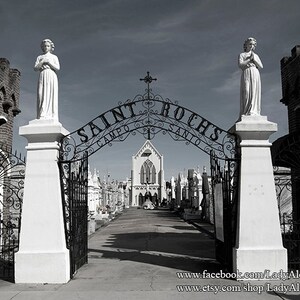 May include: A black wrought iron gate with the words "Saint Roch's Campo Santo" arched over it. Two white statues of angels stand on either side of the gate. The gate leads into a cemetery with a church in the distance.