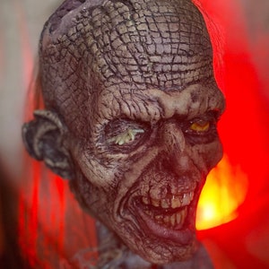 Puede incluir: Primer plano detallado de una cabeza de zombi con una superficie agrietada y texturizada. El zombi tiene ojos amarillos, dientes expuestos y una apariencia en descomposición. La cabeza está unida a una lápida que dice "FULCI LIVES".