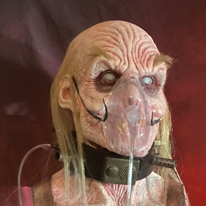 Puede incluir: Un accesorio de terror realista y perturbador que representa a una persona mayor pálida con una máscara de oxígeno médica y tubos. El accesorio lleva un collar de cuero marrón con una hebilla de metal y tiene el pelo largo y rubio. El accesorio se muestra en un soporte con dos cráneos.