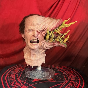 May include: A grotesque, realistic mask of a human face, half-transformed into a monstrous form. The skin is a pale pink with visible wrinkles and open mouth revealing teeth. The right side of the face is a gaping maw with yellow tendrils. The mask is mounted on a clear stand.