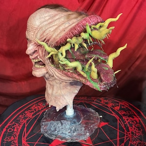 May include: A grotesque, detailed sculpture of a human head with exposed flesh and a gaping mouth. The head is mounted on a clear base. The open wound reveals a mass of green and yellow organic forms. The sculpture is set against a red backdrop.