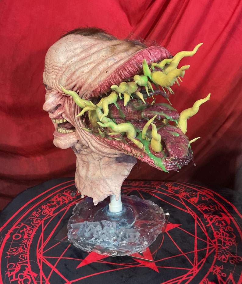 May include: A grotesque, detailed sculpture of a human head with exposed flesh and a gaping mouth. The head is mounted on a clear base. The open wound reveals a mass of green and yellow organic forms. The sculpture is set against a red backdrop.