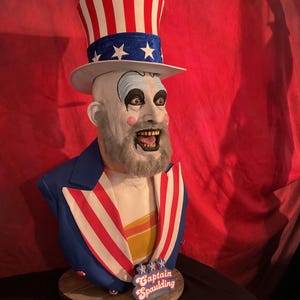 Puede incluir: Un busto de Captain Spaulding de la película House of 1000 Corpses. El busto lleva un sombrero de Tío Sam rojo, blanco y azul y una chaqueta azul con rayas rojas y blancas. El busto está sobre una base de madera con las palabras "Captain Spaulding" escritas en ella.