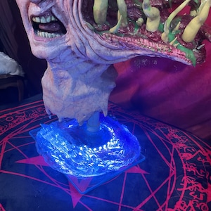 May include: A grotesque, detailed bust of a monster with exposed teeth and a gaping mouth. The sculpture is primarily flesh-toned with green and purple accents. It sits atop a clear, square base illuminated by blue LED lights, resting on a black and red patterned surface.