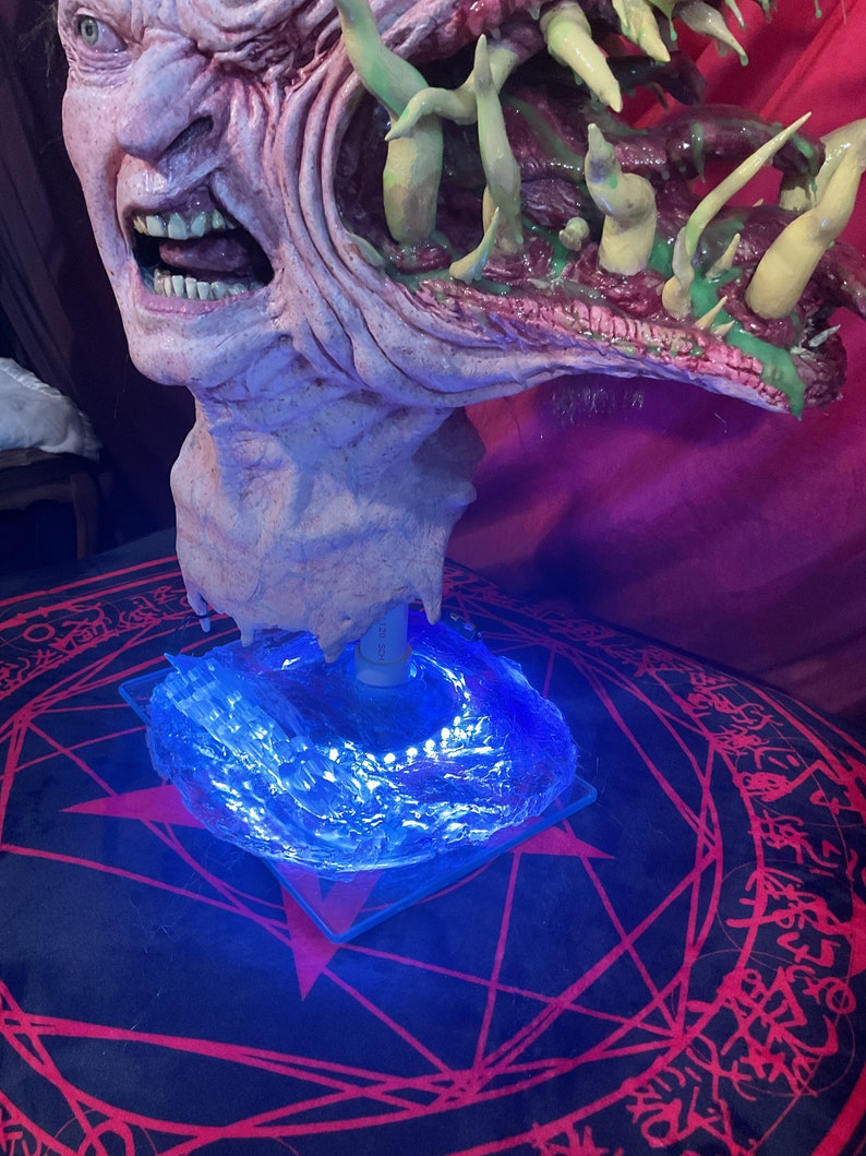 May include: A grotesque, detailed bust of a monster with exposed teeth and a gaping mouth. The sculpture is primarily flesh-toned with green and purple accents. It sits atop a clear, square base illuminated by blue LED lights, resting on a black and red patterned surface.