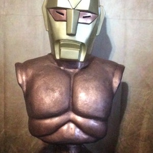 Puede incluir: Un busto de bronce de una figura que lleva un casco dorado y blanco con un solo ojo en el centro. El casco tiene una punta puntiaguda y el pecho de la figura está esculpido con músculos definidos. El busto está sentado sobre una base negra con la palabra "SPECTREMAN" escrita en letras doradas.