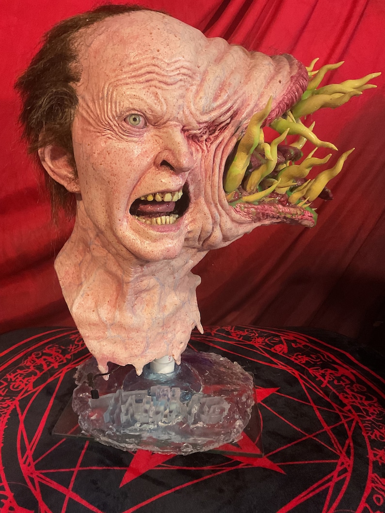 May include: A grotesque, detailed bust of a human head with a large, mutated growth on the right side. The head is pale with visible veins, wrinkles, and an open mouth. The growth features yellow, tentacle-like protrusions.