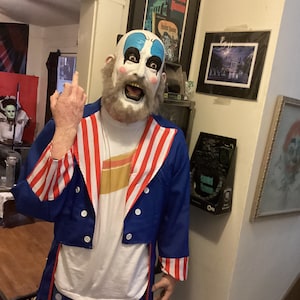 Könnte beinhalten: Eine Person in einer Clownmaske und einem Kostüm. Die Maske hat weiße Haut, blaue und rote Akzente und ein breites, zahnreiches Grinsen. Das Kostüm ist blau mit rot-weißen Streifen und weißen Punkten.