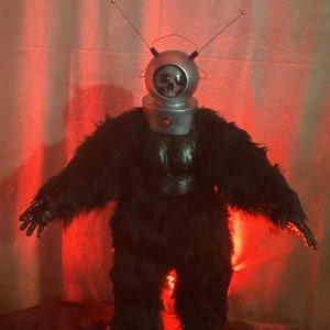 Może przedstawiać: Figurka Robot Monster z czaszką w srebrnej głowie, czarnym futrzanym ciele i anteną. Figurka ma czerwony guzik na szyi i stoi na podstawie z napisem "Robot Monster".