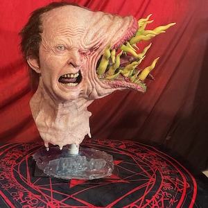 May include: A grotesque, detailed mask of a distorted human face with an open, gaping maw filled with yellow, finger-like protrusions. The mask is flesh-toned with visible wrinkles and a horrified expression. It is mounted on a clear stand.
