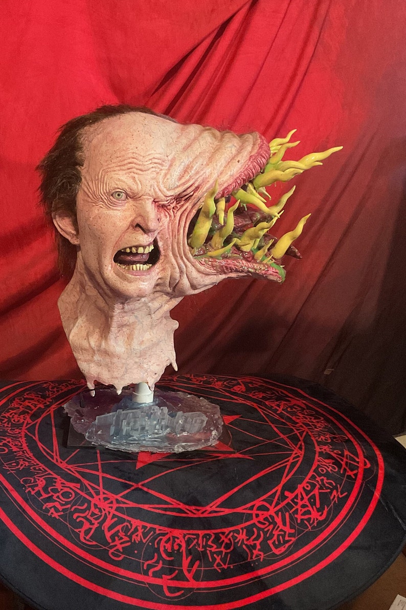 May include: A grotesque, detailed mask of a distorted human face with an open, gaping maw filled with yellow, finger-like protrusions. The mask is flesh-toned with visible wrinkles and a horrified expression. It is mounted on a clear stand.