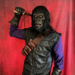 Pode incluir: Um traje do Planeta dos Macacos com uma máscara de gorila com pelo preto, um colete de couro preto e um cinto utilitário castanho. O traje inclui uma camisa roxa de manga comprida e luvas pretas. O gorila está segurando um objeto de madeira.