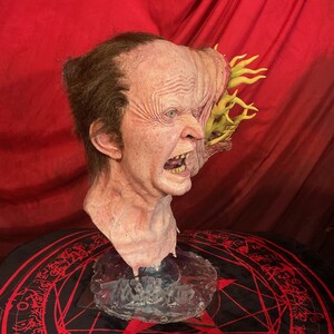 May include: A grotesque, realistic mask of a screaming, mutated face. The mask features detailed skin texture, exposed teeth, and a growth on the side with yellow tendrils. The mask is mounted on a clear base, set against a red backdrop.