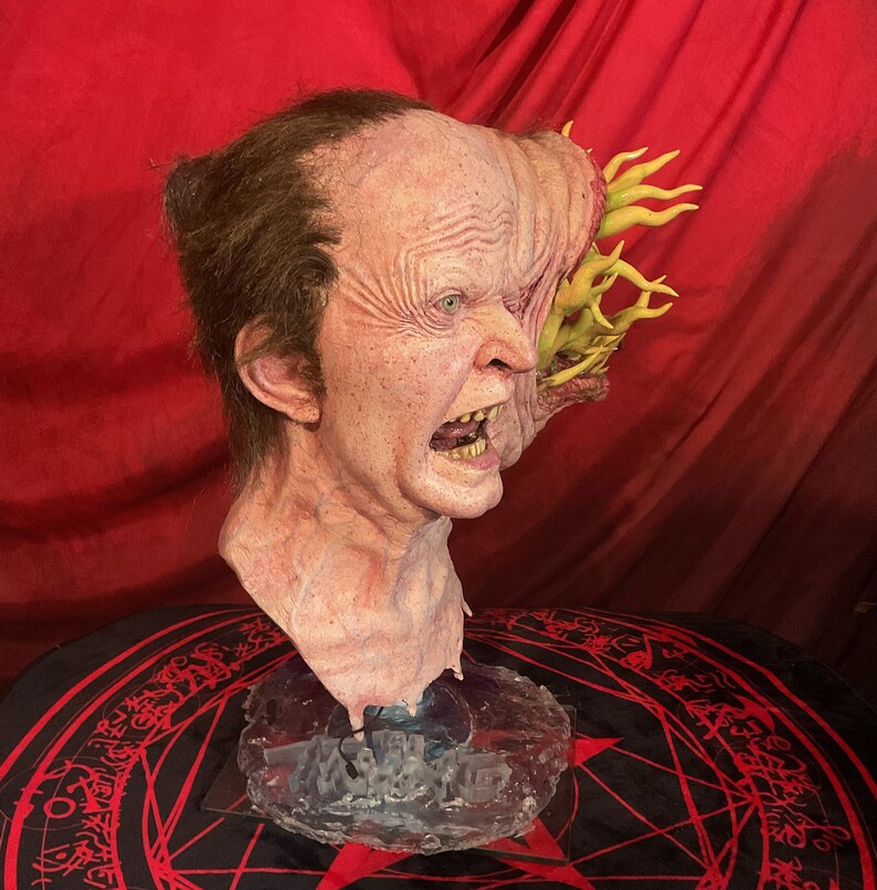 May include: A grotesque, realistic mask of a screaming, mutated face. The mask features detailed skin texture, exposed teeth, and a growth on the side with yellow tendrils. The mask is mounted on a clear base, set against a red backdrop.