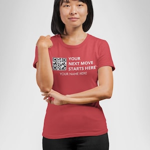 Op de afbeelding: Een rood T-shirt met de tekst "YOUR NEXT MOVE STARTS HERE" en een QR-code. Het shirt bevat ook de tekst "YOUR NAME HERE". Het model draagt een zwarte broek.