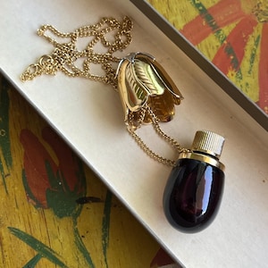 May include: A gold-tone metal necklace featuring a pendant shaped like a tulip flower with a removable cap. The pendant is attached to a dark purple, oval-shaped glass bottle with a ribbed gold-tone metal screw top, suspended by a fine gold-tone chain.