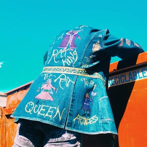 May include: A denim jacket with colorful embroidered designs and text. The jacket features illustrations of characters and the phrases "The Rat King," "Queen," and "Princess" in a whimsical font. The jacket is accented with a gold-colored trim.