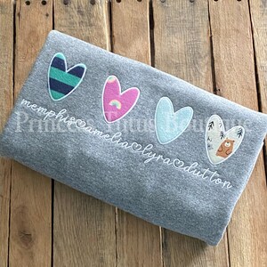 May include: A gray blanket with four heart-shaped appliqués in various colors and patterns. The blanket has the names "memphis", "amelia", "lyra", and "dutton" embroidered in white script. The hearts feature stripes, rainbows, and a bear.