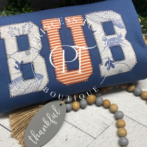 May include: A blue shirt with the word "BUB" in large, white letters with a nautical theme. The letters are outlined in blue and filled with a white and blue pattern. The "U" is orange and white striped.