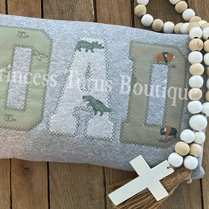 May include: A gray sweatshirt with the word "DAD" spelled out in large letters. Each letter is made of a different fabric with a dinosaur or buffalo print. A white cross with a brown tassel hangs below the sweatshirt.