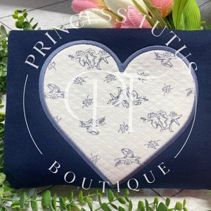 May include: A navy blue fabric heart-shaped patch with a white background and a blue floral pattern. The patch is sewn onto a navy blue fabric background.