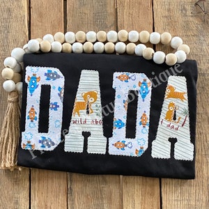May include: Black t-shirt with the word "DADA" in large, appliqued letters. The letters are decorated with rocket ships, tigers, and the phrases "wild about" and "I dad". A string of wooden beads and a tassel are in the background.