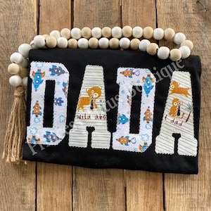 May include: Black t-shirt with the word "DADA" in white outlined letters. Each letter is made of a patchwork of fabric with a pattern of blue rockets and tigers.