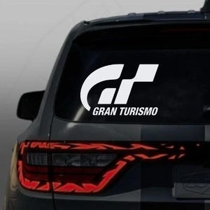 May include: White "Gran Turismo" decal on a car's rear window. The decal features a stylized "GT" logo above the words "Gran Turismo" in a sans-serif font. The car's taillights are visible below the decal.