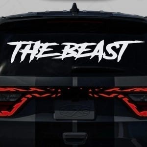 May include: White vinyl decal with the text "THE BEAST" in a bold, stylized font, applied to the rear window of a black vehicle. The vehicle's taillights are visible below the decal.