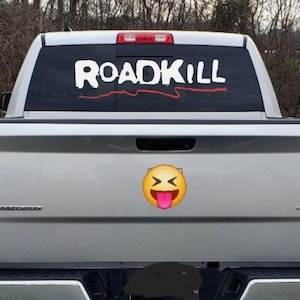 May include: A silver pickup truck with a rear window decal that reads "ROADKILL" in white letters with a red line underneath. A yellow emoji sticker with a tongue sticking out is on the truck's tailgate.
