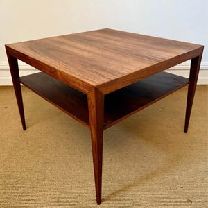May include: A square, mid-century modern side table with a dark wood finish. The table has a lower shelf and tapered legs. The top measures approximately 24 inches by 24 inches.
