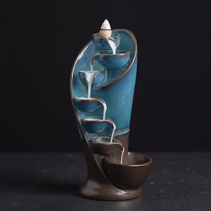 Könnte beinhalten: Ein Keramik-Räucherwasserfall in Blau- und Brauntönen. Das Design zeigt eine Reihe von kaskadierenden Schalen, mit einem weißen Rauchstrom, der nach unten fließt. Oben ist ein kegelförmiger Räucherstoff platziert.