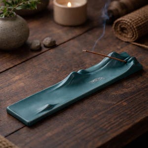 Könnte beinhalten: Ein türkisfarbener Keramik-Räucherstäbchenhalter in Form einer Bergkette. Der rechteckige Halter hat ein geschwungenes, wellenartiges Design. Ein Räucherstäbchen ist angezündet und verströmt Rauch. Eine brennende Kerze und andere dekorative Gegenstände befinden sich im Hintergrund.