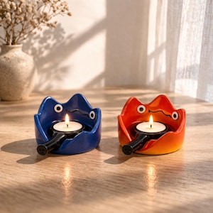Puede incluir: Dos candelabros de cerámica, uno azul y otro naranja, cada uno con forma de llama estilizada con dos círculos blancos en forma de ojos. Cada soporte contiene una vela de té encendida. Los soportes están sobre una superficie de madera.