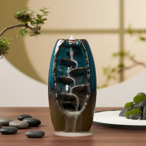 Könnte beinhalten: Ein teal-farbener und brauner Keramik-Räucherwasserfall mit Kaskadendesign. Der Räuchergefäß hat eine glänzende Oberfläche und ist wie eine Vase geformt. Weißer Rauch fließt die Ebenen hinunter und erzeugt einen visuellen Effekt. Dekorative Zen-Garten-Elemente im Hintergrund.